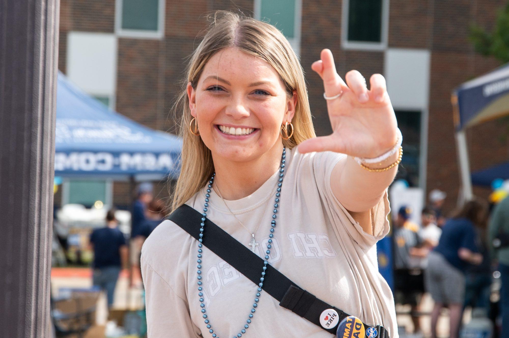 A female student wearing a light color shirt smiling at the camera, making the lions sign. In the background is other students. 